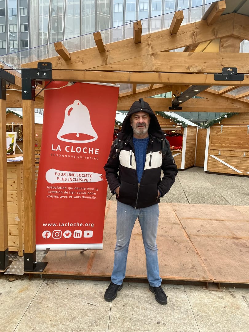 Un homme debout devant un stand de l'association La Cloche, avec un panneau rouge mettant en avant la mission de solidarité sociale et d'inclusion.